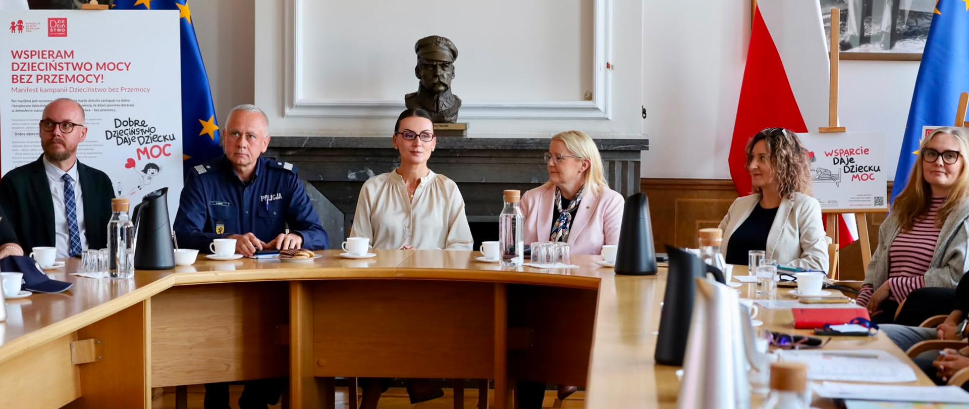 Grupa osób siedzi przy dużym drewnianym stole podczas spotkania w sali urzędowej. Wśród uczestników znajduje się umundurowany policjant. W tle stoją flagi Polski i Unii Europejskiej oraz plakaty kampanii „Dzieciństwo bez przemocy” z hasłem „Wsparcie daje dziecku moc”.