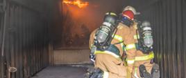 Strażacy klęcząc w komorze gaszą symulowany pożar. Na suficie palą się przygotowane materiały palne.
