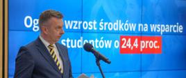 Zdjęcie z boku, minister Wieczorek stoi na sali, trzyma w ręku kartkę papieru i mówi do mikrofonu na stojaku, za nim na wbudowanym w ścianę wielkim ekranie napis Ogólny wzrost środków na wsparcie studentów o 24,4 proc.