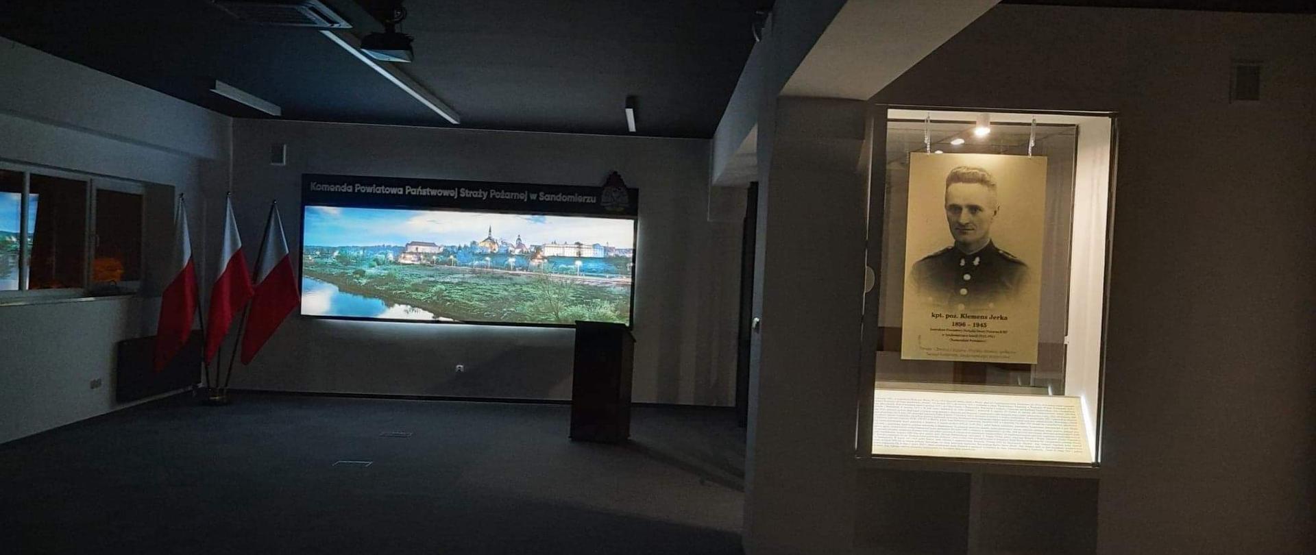 Zdjęcie zrobione w ciemnej reprezentacyjnej sali. Od lewej trzy flagi Rzeczpospolitej Polskiej, następnie podświetlona panorama Sandomierz i z prawej strony podświetlona gablota ze zdjęciem kapitana Klemensa Jerki i jego notą biograficzną.