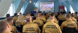 Na sali, w rzędach siedzą oficerowie PSP - ok. 30 osób, w pierwszych rzędach goście narady. Widok na stół prezydialny i ekran, gdzie w ramach transmisji wideo rozmówcą jest zastępca komendanta głównego. Strażacy w mundurach służbowych - musztardowych z czarnym napisem Paśńtwowa Straż Pożarna