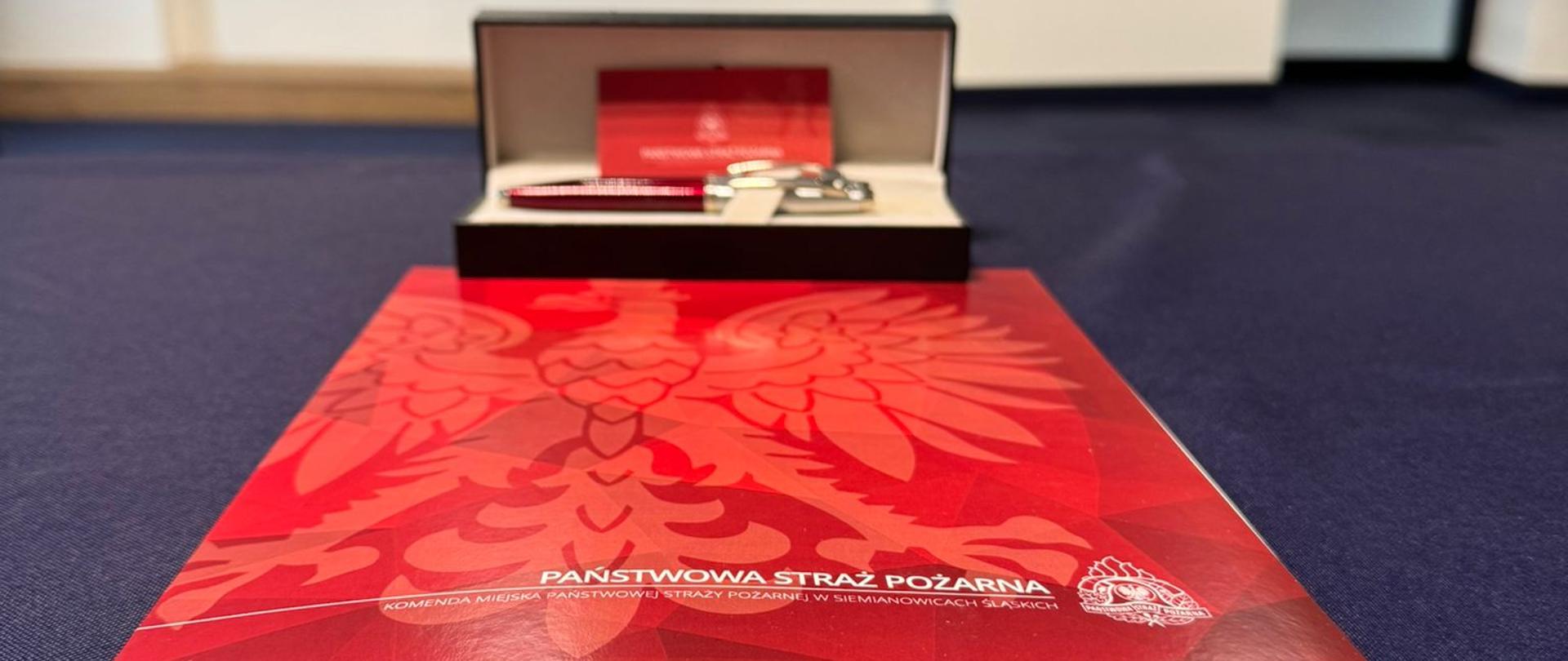 Akt ślubowania leżący na stole przykrytym granatowym suknem, za nim pudełko z długopisami oraz wizytówką z logiem PSP.