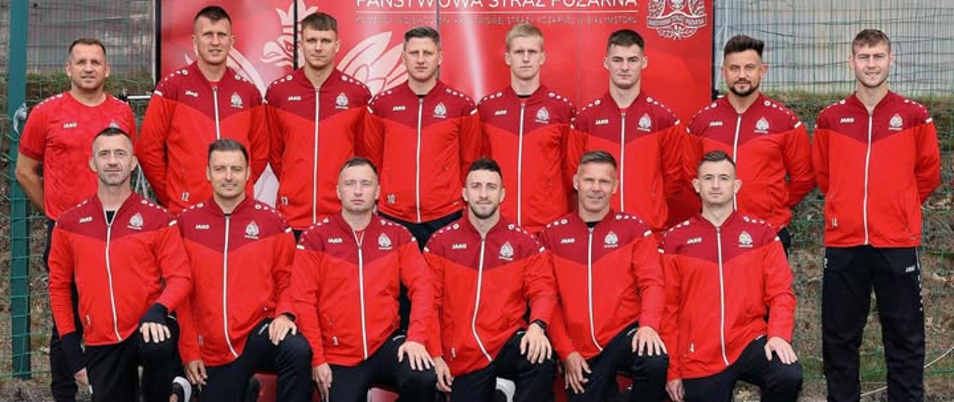 Na zdjęciu znajduje się drużyna piłkarska strażaków. Zawodnicy mają na sobie jednakowe stroje sportowe: czerwone bluzy z bordowym górnym panelem i czarne spodnie. Wszyscy stoją lub klęczą na sztucznej murawie boiska. Grupa składa się z czternastu mężczyzn, ośmiu stoi w tylnym rzędzie sześciu klęczy lub siedzi w rzędzie przednim. Za nimi widoczna jest czerwona tablica z logotypem i napisem: „Państwowa Straż Pożarna – Komenda Wojewódzka Państwowej Straży Pożarnej w Białymstoku”. Na dole zdjęcia znajdują się dwa logotypy i napis: „IV Mistrzostwa Polski Strażaków w Piłce Nożnej 6-osobowej 17-18.09.2025 Białystok”. Na trawie, przed drużyną, leży piłka nożna.