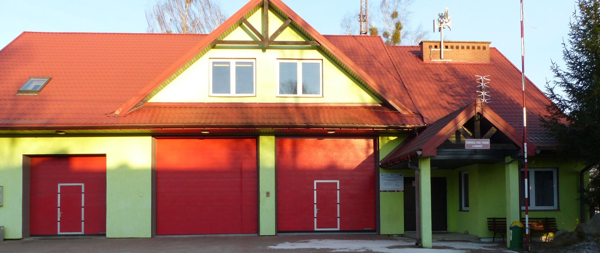 Remiza OSP Białowieża. Dwa duże wjazdy garażowe z bramami czerwonymi, dodatkowo po prawej stronie mniejsza czerwona brama, widoczne po lewej stronie wejście główne do budynku z poddaszem użytkowym, elewacja żółta z czerwonym dachem, widoczny maszt biało-czerwony oraz maszt antenowy, podjazd utwardzony kostka brukową. 