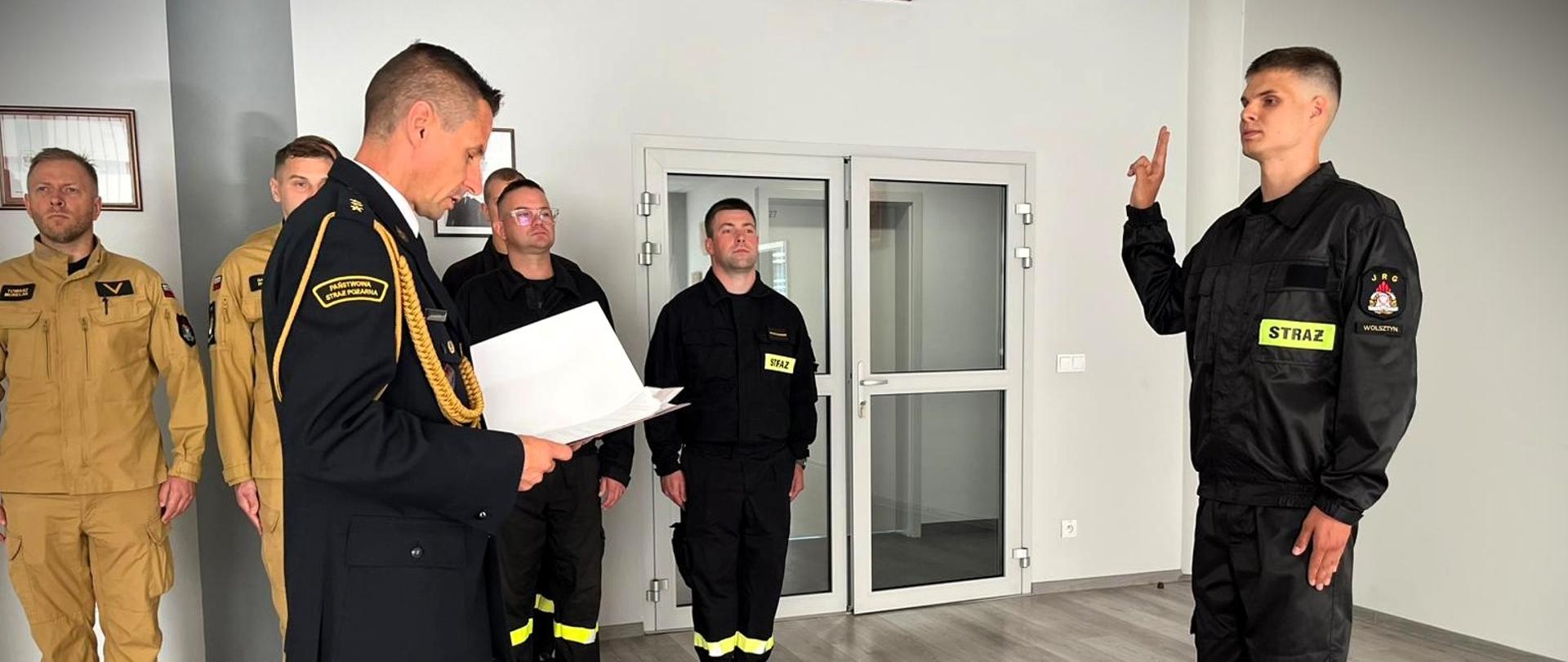 Na zdjęciu widzimy uroczysty moment w siedzibie Komendy Powiatowej Państwowej Straży Pożarnej w Wolsztynie.
Po prawej stronie kadru stoi młody mężczyzna w ubraniu koszarowym. Jego postawa jest poważna i formalna.
Po lewej stronie, stoi strażak w galowym mundurze i czyta dokument – akt ślubowania.
Całość ma bardzo oficjalny i patriotyczny charakter – obecne są godło Polski, barwy narodowe oraz emblematy straży.
