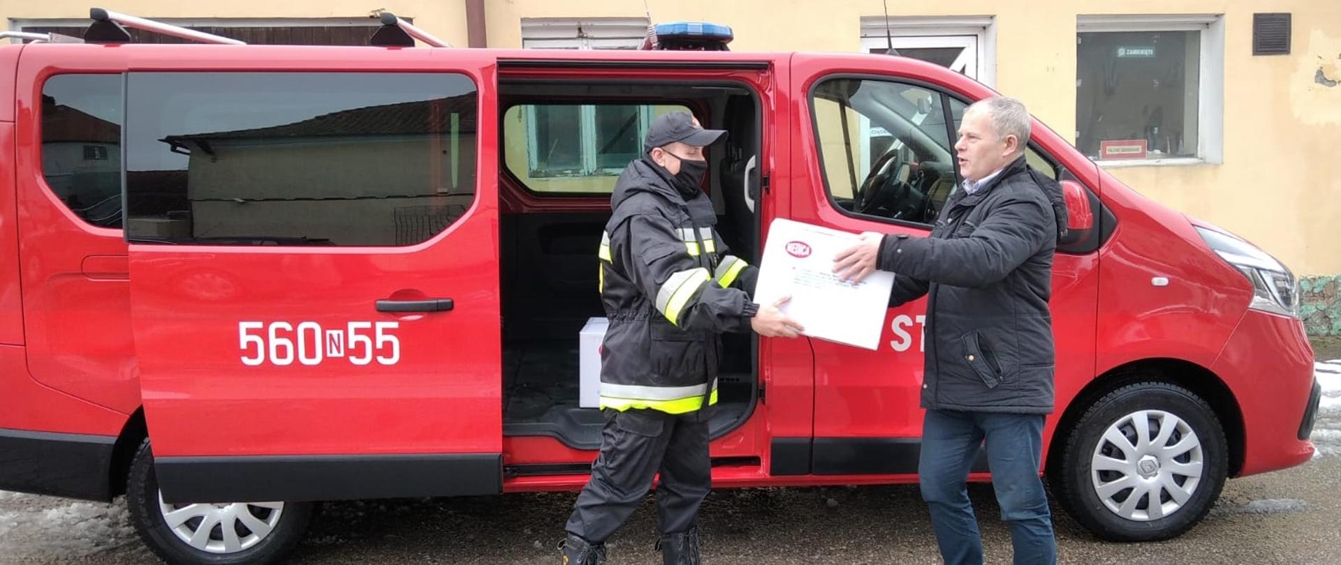 Strażak przekazuje karton dla urzędnika w którym znajdują się maseczki medyczne. Za nimi znajduje się czerwony samochód PSP bus z otwartymi bocznymi drzwiami. 