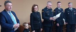 Zdjęcie obrazuje grono zaproszonych gości, pośród nich stoi Komendant Powiatowy który to odczytuje zapisy w decyzji o przyznaniu świadczenia ratowniczego. 