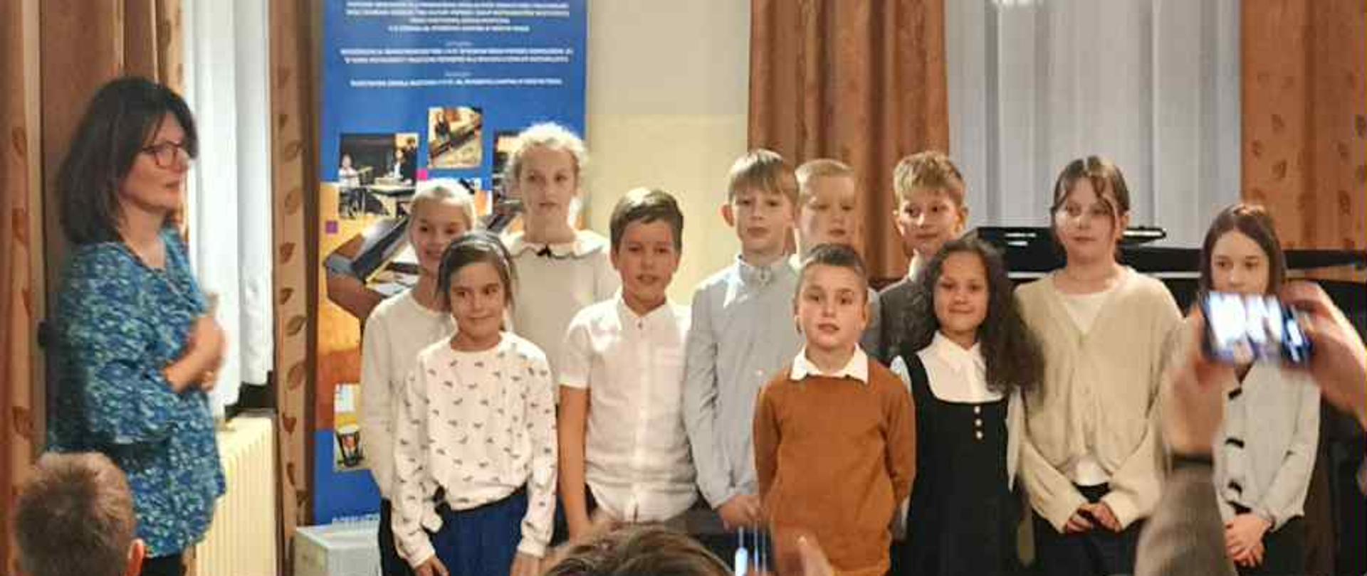 Zdjęcie przedstawia grupę dzieci, prawdopodobnie uczniów szkoły muzycznej po występie, oraz kilka osób dorosłych w jasnym, klasycznie urządzonym pomieszczeniu.W Centrum i Po Prawej: Stoi dwanaścioro dzieci (dziewczynki i chłopcy), ustawionych w dwóch rzędach. Ubrane są w eleganckie, jasne stroje, przeważnie białe, beżowe i brązowe, co sugeruje formalną okazję lub koncert. Wyróżnia się dziewczynka w ciemnej (granatowej lub czarnej) sukience. Patrzą w kierunku aparatu i wyglądają na zadowolone lub dumne.
Po Lewej: Stoi dorosła kobieta- nauczycielka w niebieskiej bluzce z wzorem, uśmiecha się i patrzy na dzieci. Na Pierwszym Planie (Tyłem): Znajdują się dwie osoby dorosłe. Jedna (głównie widoczne plecy) jest odwrócona do widza i wykonuje gest klaskania w kierunku dzieci. Druga (po prawej) trzyma aparat lub telefon i robi zdjęcie stojącym dzieciom. Wnętrze: Jest to pokój o neutralnym, jasnym kolorze ścian. Na podłodze widoczne są krzesła kinowe (czarne, składane), ustawione w rzędach, co sugeruje, że odbyła się tu widownia lub wystąpienie.
Oświetlenie: Z sufitu zwisają trzy długie, czarne kable z klasycznymi, białymi kloszami lamp. Okna i Dekoracje: W tle, po obu stronach ściany, znajdują się duże okna zasłonięte ciężkimi, brązowymi (w odcieniu miedzi) zasłonami i firankami. Między oknami widać baner lub plakat z niebieskimi elementami i napisami, prawdopodobnie informujący o instytucji lub wydarzeniu. Ogólne wrażenie: Zdjęcie ma ciepły i radosny charakter, uwieczniając moment podziękowania i pamiątkową chwilę po zakończeniu wydarzenia muzycznego.