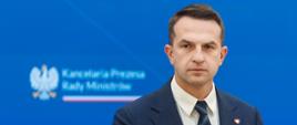 Rzecznik rządu, Minister Adam Szłapka podczas konferencji prasowej 24.06.2025 r. W tle ekran Kancelarii Prezesa Rady Ministrów oraz media społecznościowe rzecznika rządu. Obok flagi Polski i Unii Europejskiej.