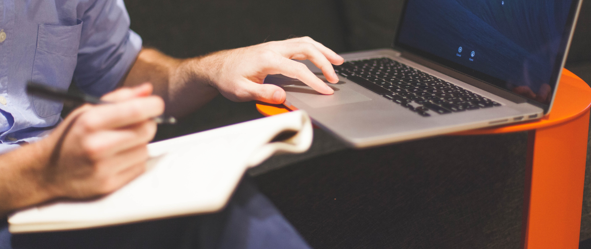 Siedzący mężczyzna w niebieskiej koszuli, ujęcie od połowy ramienia. Na kolanie trzyma notatnik, opiera o niego prawą dłoń z długopisem. Lewą dłoń trzyma na touchpadzie laptopa, który stoi na pomarańczowym stoliku. 