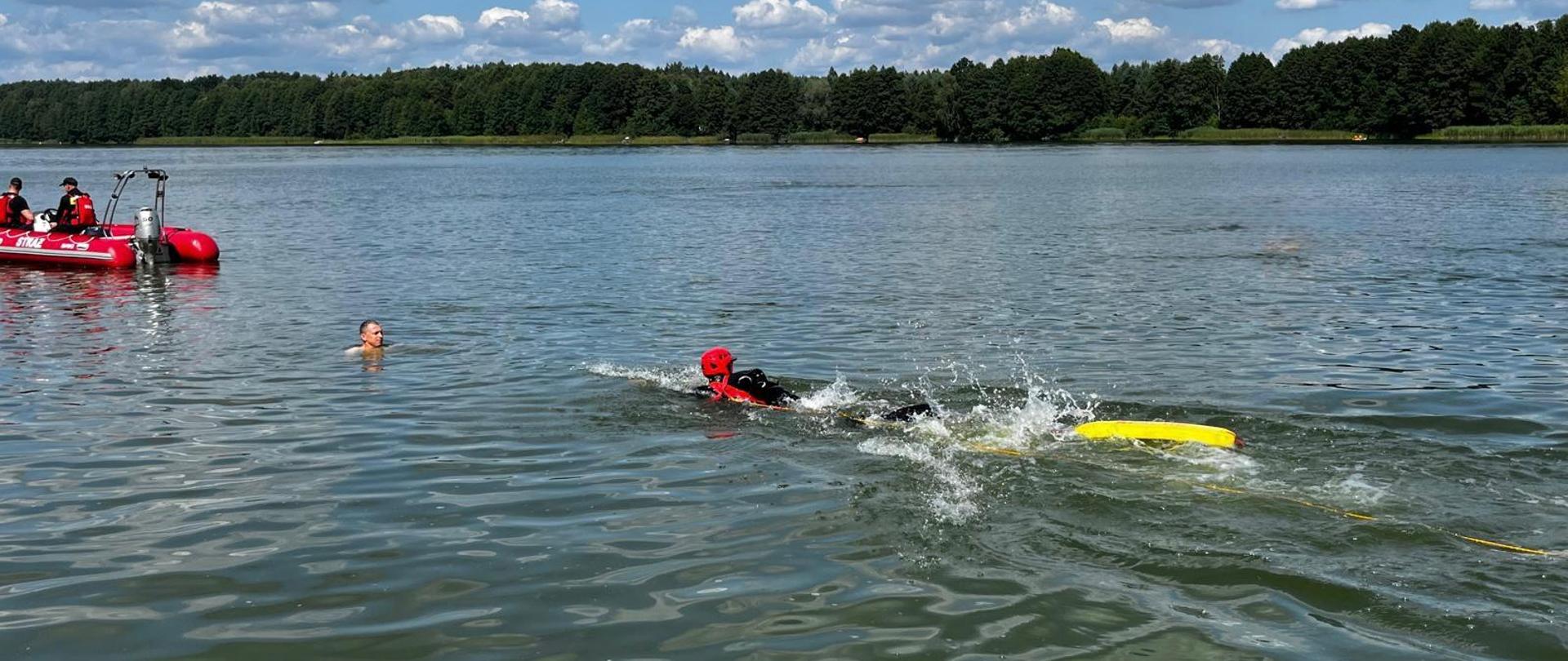 Ratownik w czerwonym kasku i czarnej piance płynie z żółtą bojką ratunkową w kierunku osoby znajdującej się w wodzie. W tle po lewej stronie stoi łódź z ratownikami. Pogoda jest słoneczna, na niebie rozproszone białe chmury.