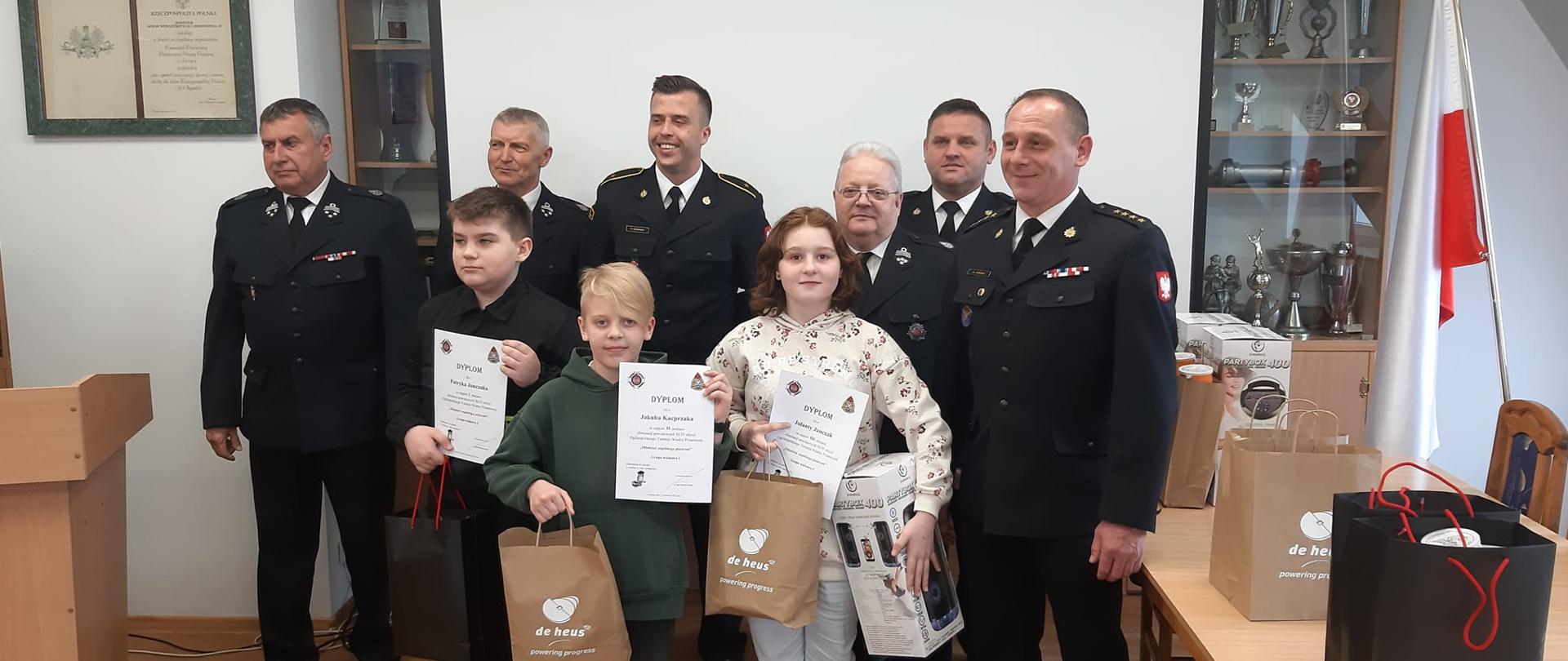 Na zdjęciu widać dziewięć osób, sześciu strażaków i troje dzieci. Trzech strażaków ubranych jest w mundur Państwowej Straży Pożarnej a trzech w mundur ochotniczej straży pożarnej. Wszyscy pozują do zdjęcia. Dzieci trzymają w rękach torebki z nagrodami i dyplomy. W tle rozwinięty jest biały ekran do rzutnika. Z boku na stołach stoją nagrody i torebki. W tle widać flagę Polski. Zdjęcie zrobione jest w pomieszczeniu.