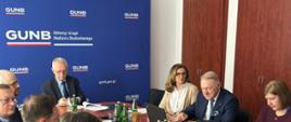 Posiedzenie Rady Wyrobów Budowlanych w Głównym Urzędzie Nadzoru Budowlanego. Uczestnicy spotkania siedzą przy stole, korzystając z laptopów i dokumentów. W tle widoczne jest logo GUNB, a na stole znajdują się butelki wody i inne akcesoria. 