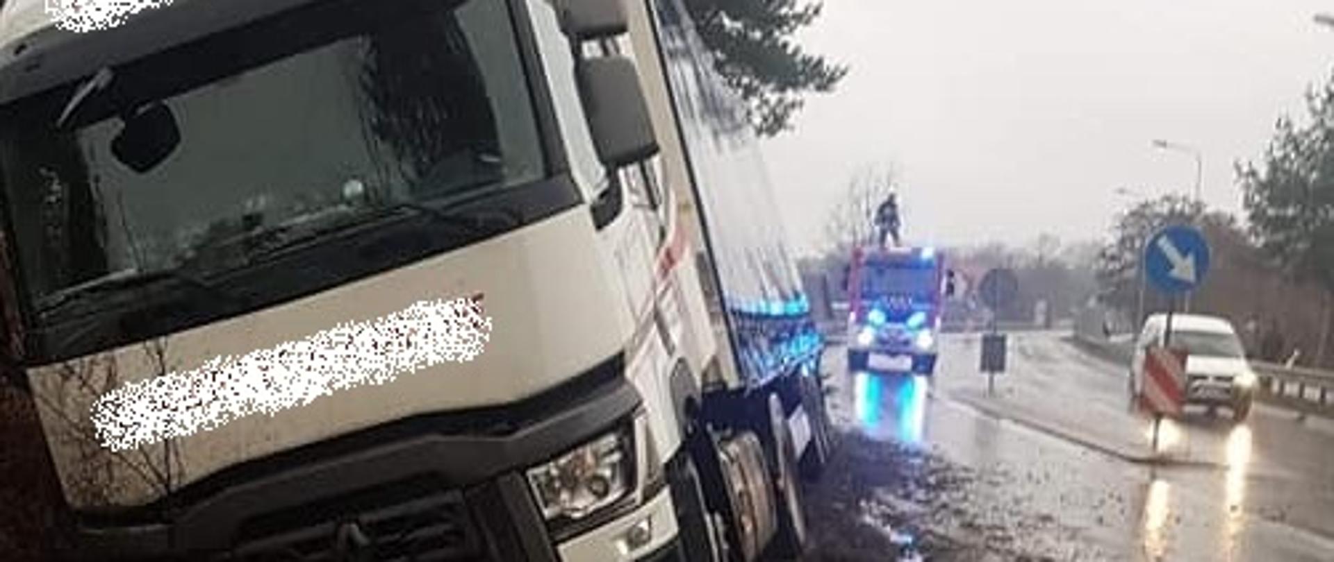 Samochód ciężarowy w rowie, w oddali widać samochód straży pożarnej, obok jedzie biały samochód osobowy