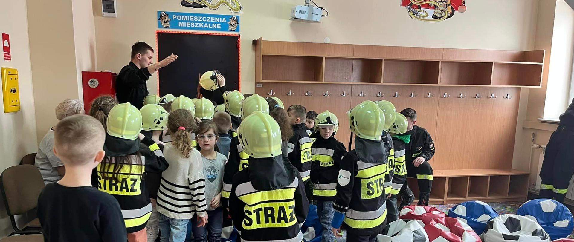 Zdjęcie przedstawia dzieci na Sali edukacyjnej, niektóre ubrane w specjalne ubrania na wzór straży pożarnej. Strażak w umundurowaniu koszarowym objaśnia dzieciom tematykę kształtowania prawidłowych zachowań i nawyków w sytuacji zagrożenia życia i zdrowia.