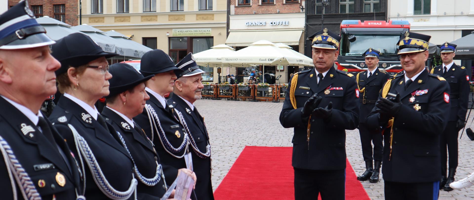 W szeregu po lewej stronie stoją druhowie i druhny OSP, po prawo na czerwonym dywanie Zastępca komendanta głównego PSP wraz z komendantem wojewódzkim PSP 