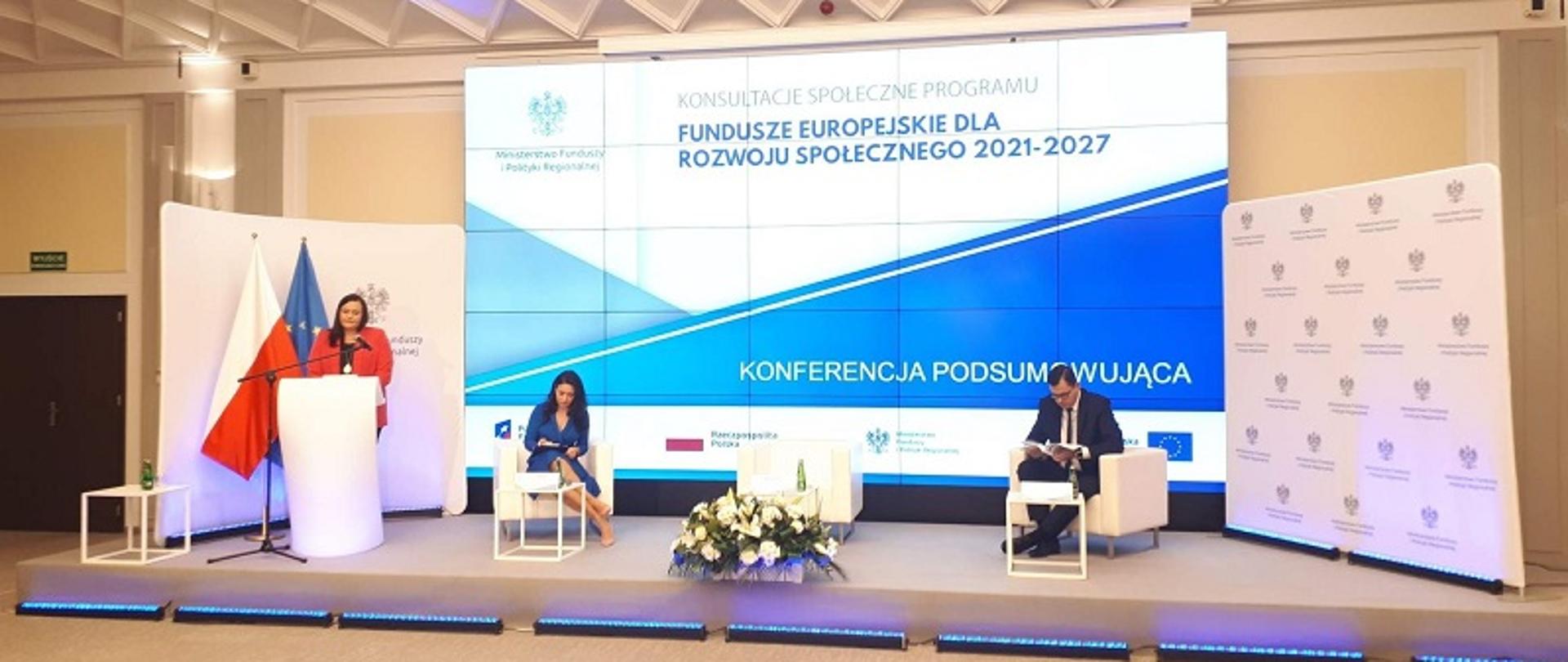 Sala konferencyjna. Minister Jarosińska-Jedynak stoi przy mównicy, za nią flagi Polski i UE oraz ekran z napisem Fundusze Europejskie dla Rozwoju Społecznego 2021-2027 - konferencja podsumowująca. Obok minister siedzą kobieta i mężczyzna.