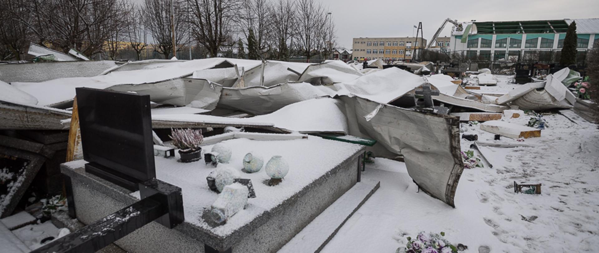 Zdjęcie wykonane na cmentarzu. Na pierwszym planie widoczne uszkodzone zerwanym dachem nagrobki. Na drugim planie widać uszkodzony budynek hali gimnastycznej.
