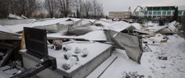 Zdjęcie wykonane na cmentarzu. Na pierwszym planie widoczne uszkodzone zerwanym dachem nagrobki. Na drugim planie widać uszkodzony budynek hali gimnastycznej.