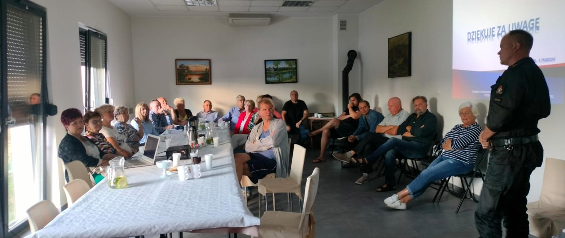 Uczestnicy prelekcji siedzący przy stole. Strażak prowadzący pogadankę stoi bokiem.