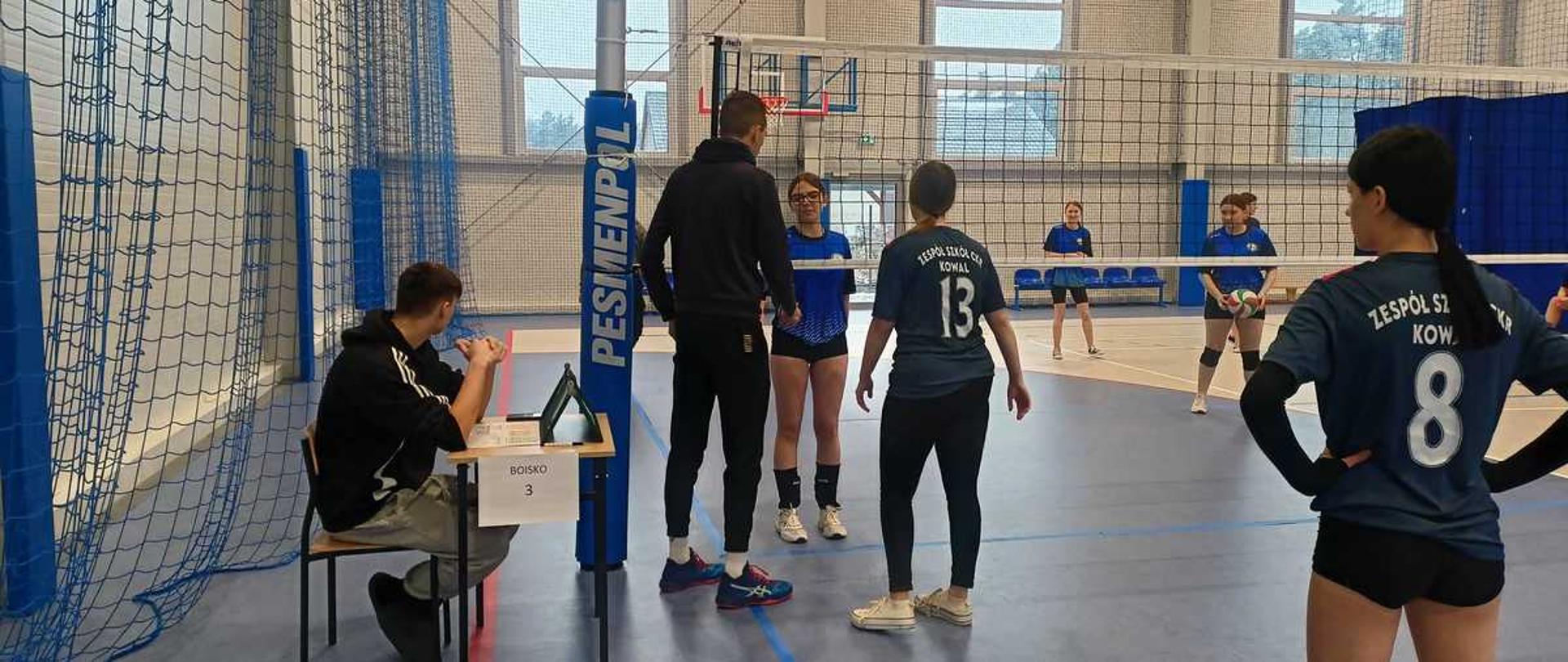 Zdjęcie przedstawia scenę z meczu siatkówki, prawdopodobnie rozgrywanego w hali sportowej należącej do Zespołu Szkół w Kowalu. Na boisku widoczne są zawodniczki i zawodnicy w strojach sportowych.
Opis szczegółowy
Pierwszy plan: Po lewej stronie, za siatką, siedzi osoba przy stoliku sędziowskim, skupiona na tablecie lub karcie wyników. W centrum, na środku boiska, stoi chłopak w czarnej bluzie i spodniach dresowych oraz dziewczyna tyłem do obiektywu, w ciemnym T-shircie z białym numerem "13" i napisem "ZESPÓŁ SZKÓŁ KOWAL".
Tło: Po prawej stronie kadru stoi inna zawodniczka tyłem, również w ciemnym stroju z białym numerem ""8"". Inni gracze w niebieskich koszulkach są widoczni w tle, za siatką.
Hala: Hala jest dobrze oświetlona, z drewnianym parkietem i jasnymi ścianami. Widoczny jest sprzęt sportowy, w tym słupek do siatkówki z niebieską osłoną z napisem "PESMENPOL".
Atmosfera: Scena ukazuje moment koncentracji i rywalizacji podczas wydarzenia sportowego.