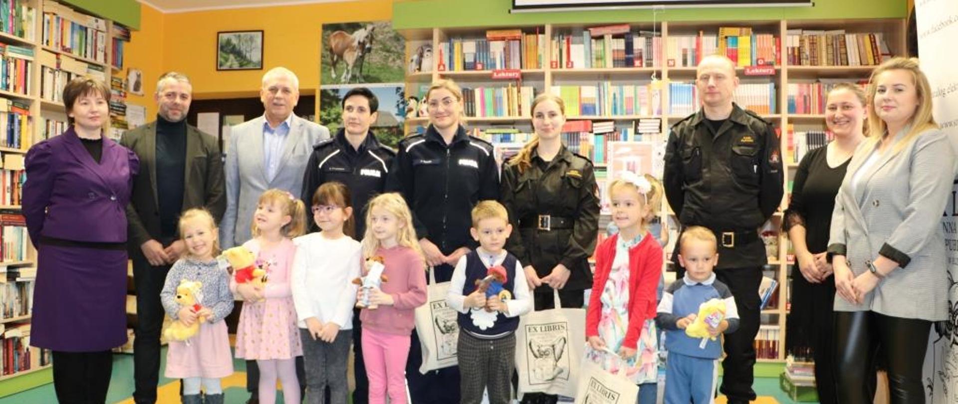 Zdjęcie wykonane wewnątrz biblioteki. Oświetlenie sztuczne. Widać funkcjonariuszy, pracowników biblioteki oraz dzieci, które brały udział w konkursie.