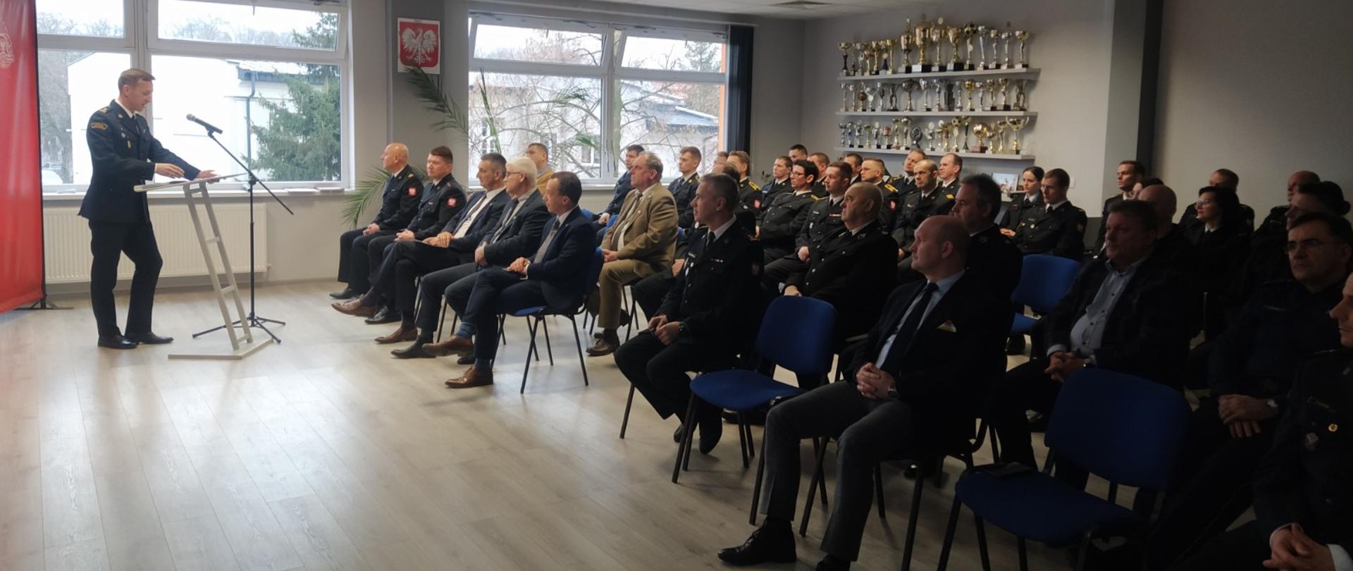 Zdjęcie przedstawia Zastępcę Wielkopolskiego Komendanta Wojewódzkiego PSP podczas przemowy na naradzie rocznej. Osoba stoi przy mównicy. Zdjęcie zrobione na sali. W tle widać baner z napisem "Państwowa Straż Pożarna". Na krzesłach siedzą zaproszeni goście.