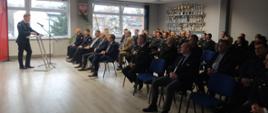 Zdjęcie przedstawia Zastępcę Wielkopolskiego Komendanta Wojewódzkiego PSP podczas przemowy na naradzie rocznej. Osoba stoi przy mównicy. Zdjęcie zrobione na sali. W tle widać baner z napisem "Państwowa Straż Pożarna". Na krzesłach siedzą zaproszeni goście.