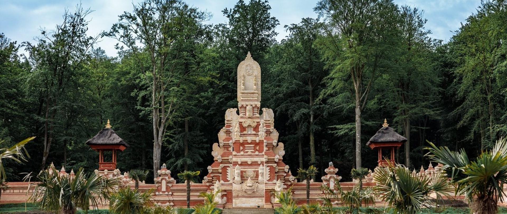 Widok na świątynię Vishnu, do której prowadzą szerokie kamienne schody. Budowlę otaczają palmy i czerwone ceremonialne parasole, a jej wejście ozdobiono biało-żółtą tkaniną.
