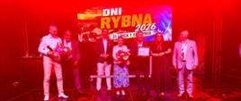 Grupa uczestników Dni Rybna 2026 stoi na scenie, oświetlona czerwonym światłem. Wśród osób znajdujących się na scenie jest wicewojewoda.