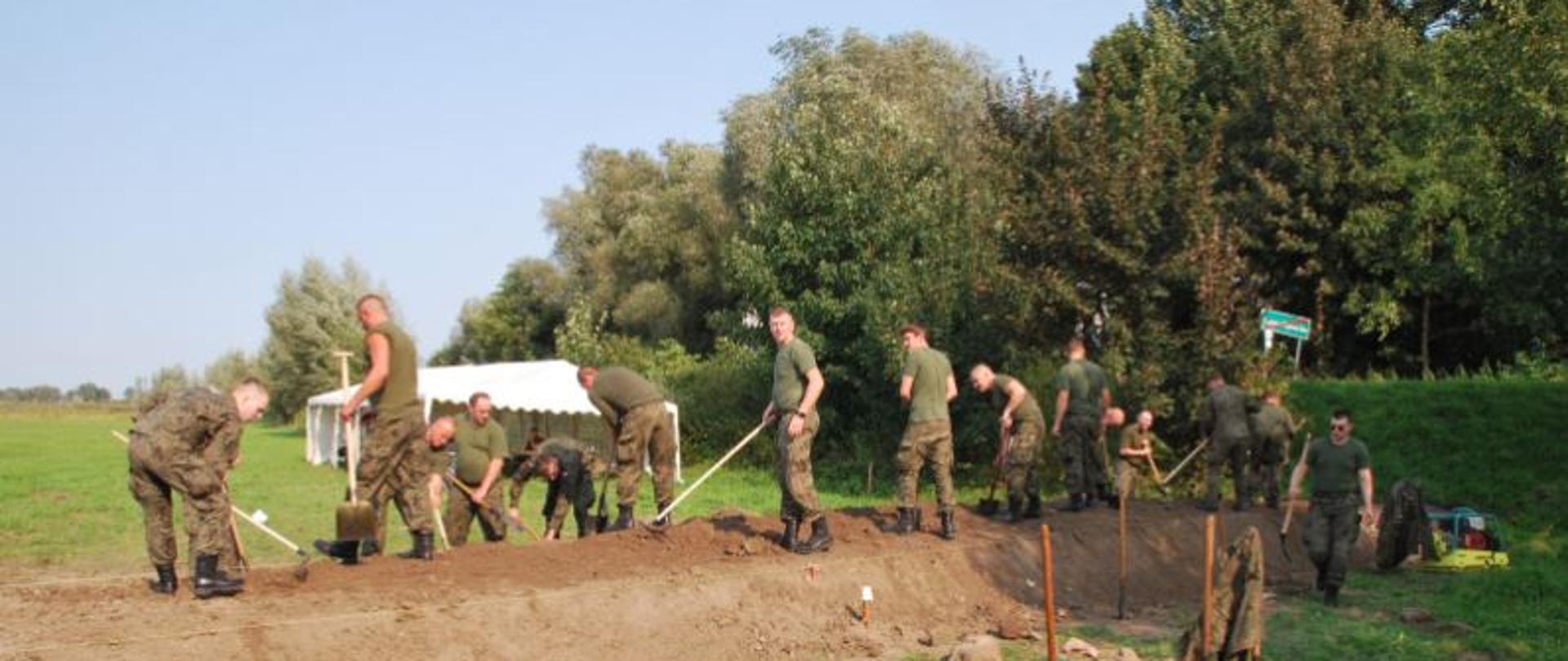 Zdjęcie przedstawia żołnierzy Wojsk Obrony Terytorialnej podczas budowy wału. Żołnierze stoją w rzędzie u uklepują przy użyciu łopat wysoki na metr, usypany z ziemi wał.