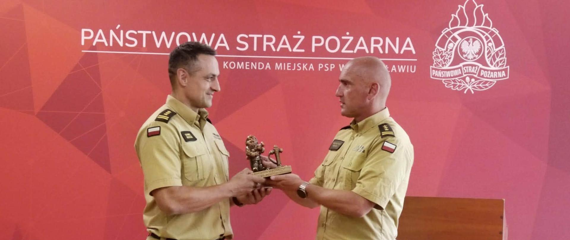 Dwóch strażaków Państwowej Straży Pożarnej w jasnych mundurach stoi naprzeciw siebie podczas uroczystości i wspólnie trzyma pamiątkową statuetkę przekazywaną z rąk do rąk. W tle znajduje się czerwona ścianka z napisem „Państwowa Straż Pożarna – Komenda Miejska PSP we Wrocławiu” oraz godłem PSP. Po prawej stronie widoczna jest mównica z godłem Rzeczypospolitej Polskiej.