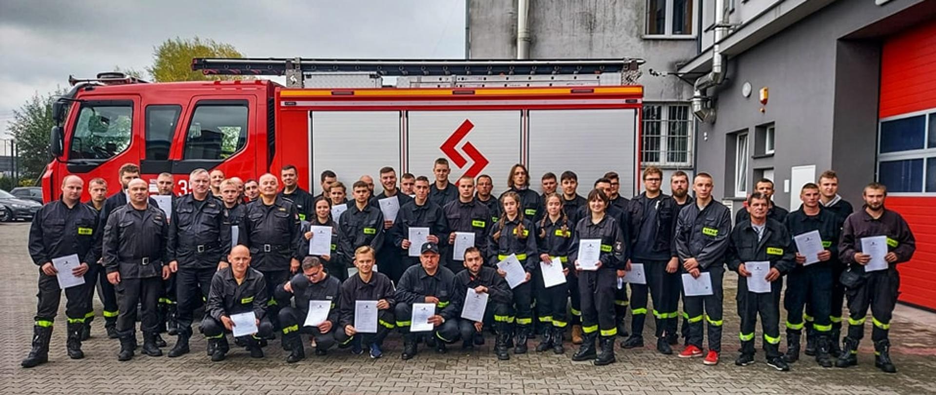 Zdjęcie przedstawia grupę strażaków trzymających certyfikaty ubranych w ubrania koszarowe koloru czarnego na tle pojazdu strażackiego koloru czerwonego i budynku.
