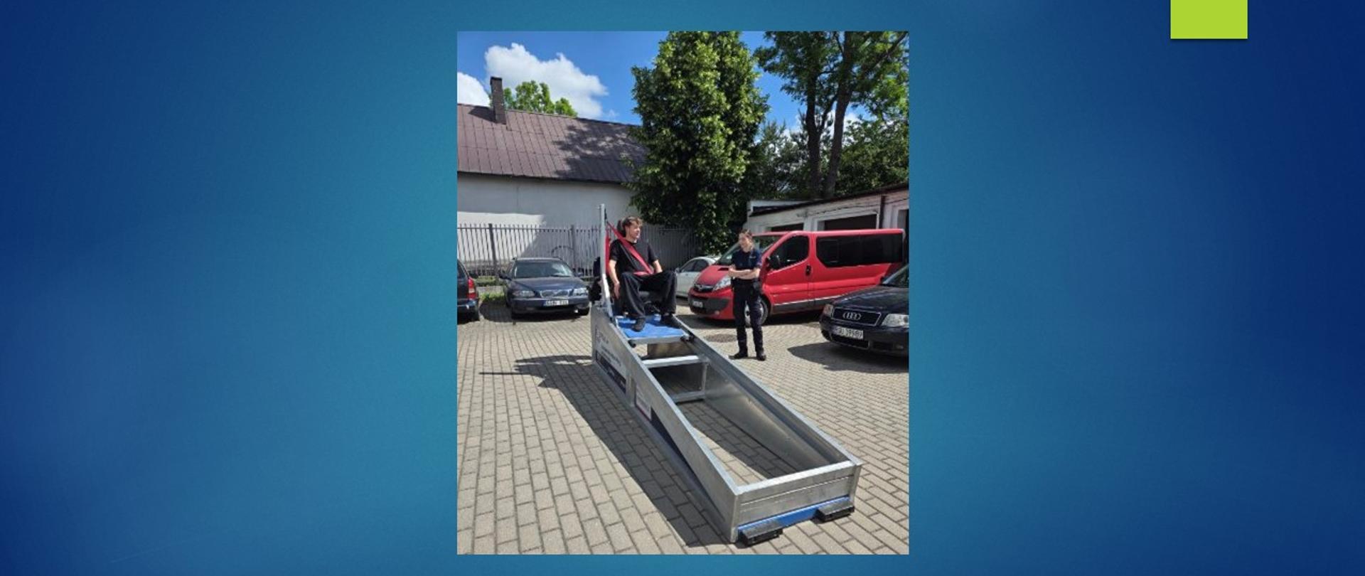 Młody chłopak siedzi na symulatorze zderzeń, ma zapięte pasy bezpieczeństwa, obok stoi policjantka.