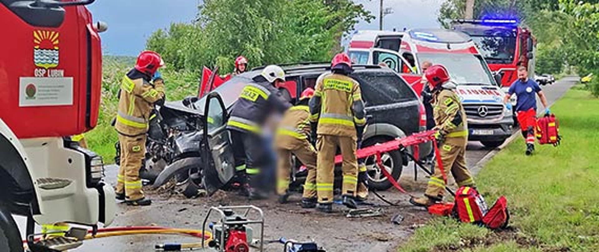Zdjęcie przedstawia wydostanie poszkodowanej osoby przez strażaków z rozbitego pojazdu który stoi na drodze.