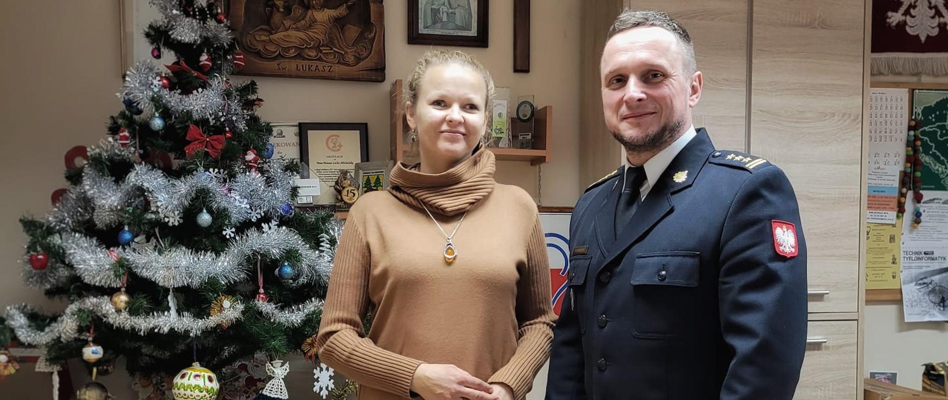Na tle choinki stoi kobieta oraz mężczyzna w mundurze.