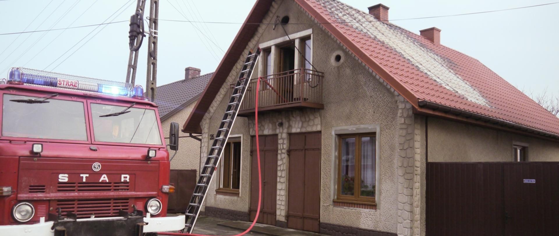 Po lewej stroni stoi samochód pożarniczy. W centralnej części o balkon budynku oparta drabina i przeciągnięty waż. Przebarwiona blacha od temperatury.