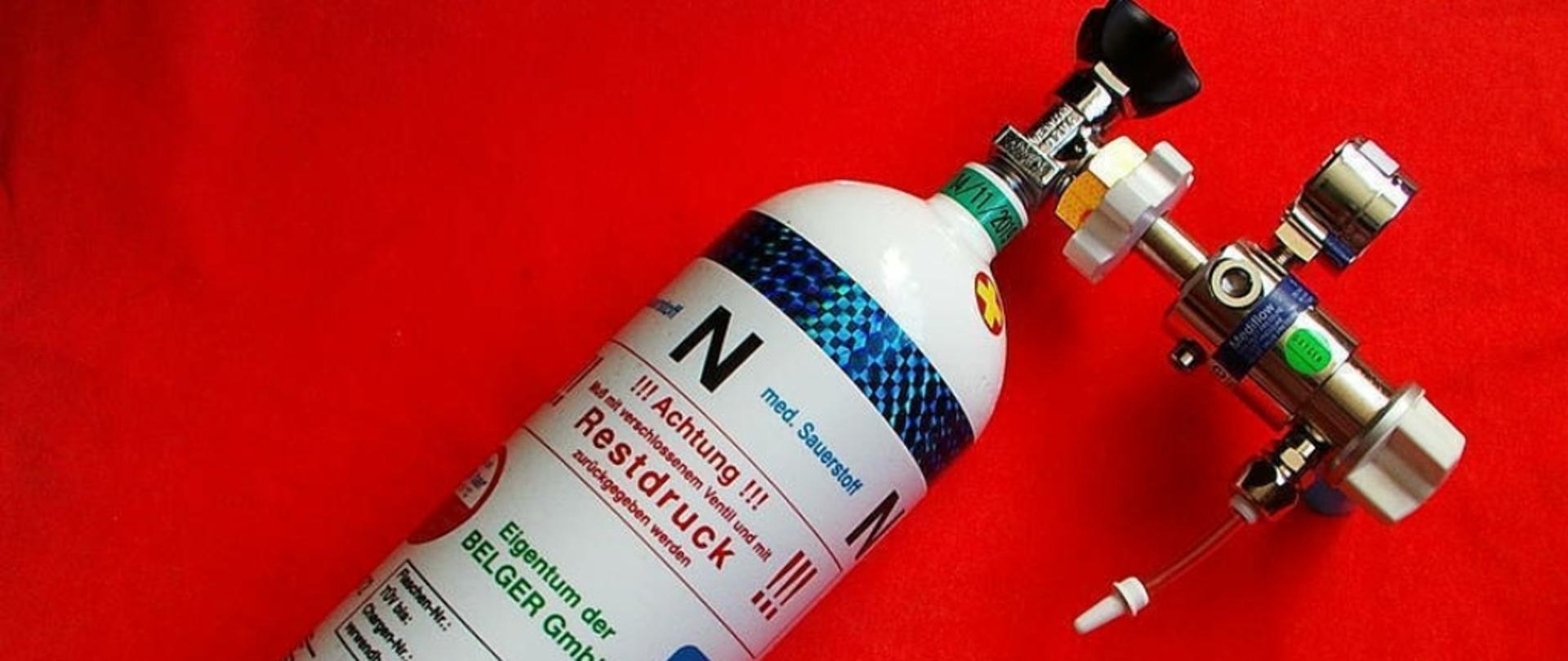 Na zdjęciu butla tlenowa koloru białego z naklejonymi oznaczeniami oraz podłączony do niej reduktor 