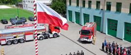 Widok z góry na plac przed remizą strażacką. Na pierwszym planie znajduje się wysoki maszt z dużą biało-czerwoną flagą Polski. Na placu stoją dwa czerwone wozy strażackie oraz ustawieni w szyku strażacy w galowych mundurach i poczty sztandarowe. W tle widoczny jest budynek straży pożarnej z kilkoma bramami garażowymi oraz otaczająca go zieleń. Zdjęcie przedstawia monitor komputera wyświetlający fotografię z uroczystości strażackiej