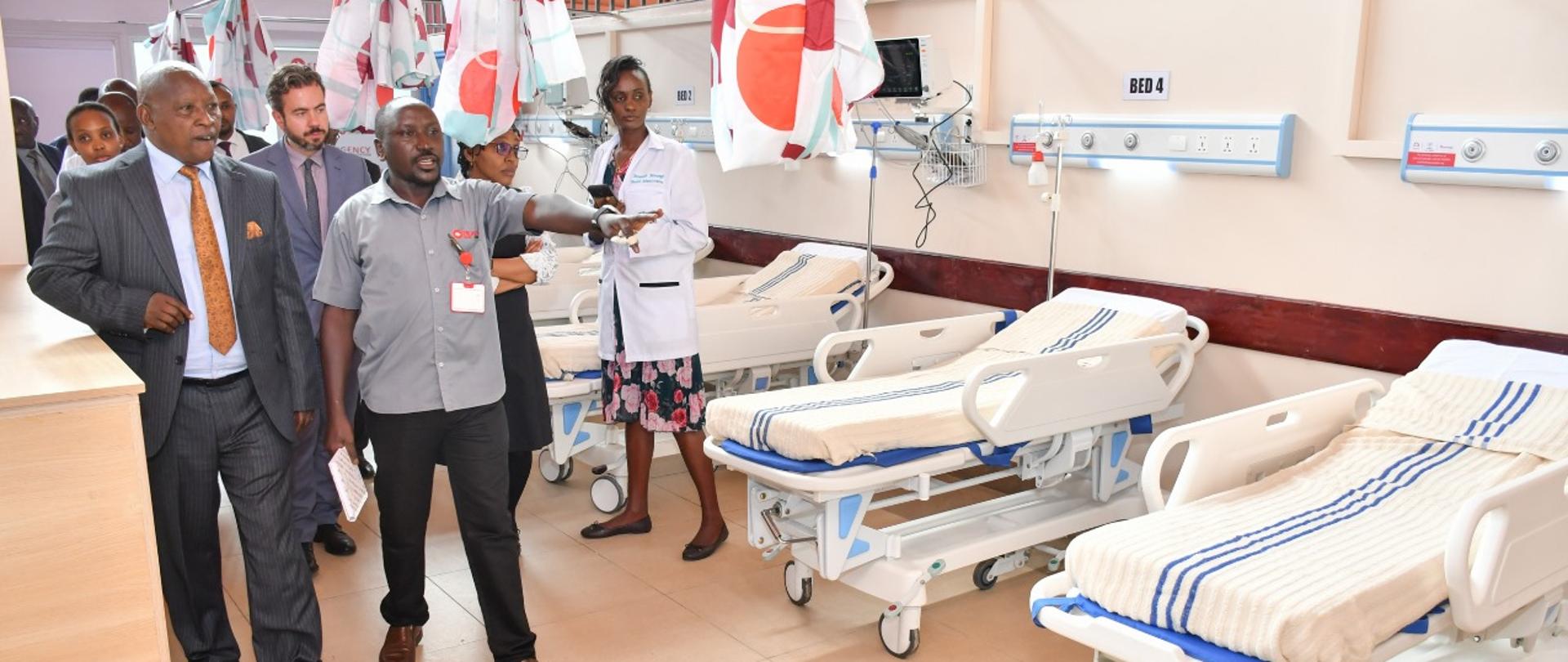 Grupa osób odwiedza oddział szpitalny, widać łóżka, jeden mężczyzna pokazuje i wyjaśnia coś grupie osób idącej za nim