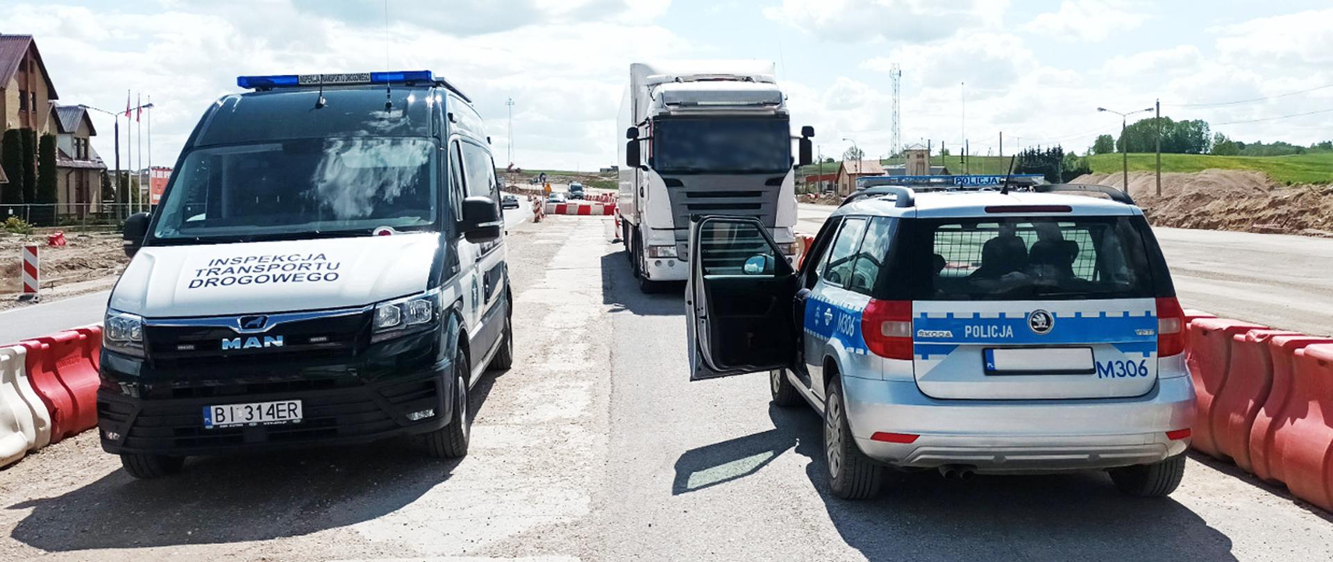 Zespół pojazdów zatrzymany do kontroli przez inspektorów podlaskiej Inspekcji Transportu Drogowego. Na pierwszym planie inspekcyjny furgon i policyjny radiowóz. Za nim skontrolowana ciężarówka. W tle ruch na drodze.