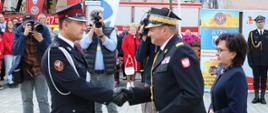 Zastępca komendanta głównego PSP podaje dłoń strażakowi w geście gratulacji, obok komendanta stoi pani marszałek, w tle uczestnicy wydarzenia i dwóch mężczyzn z aparatami fot.