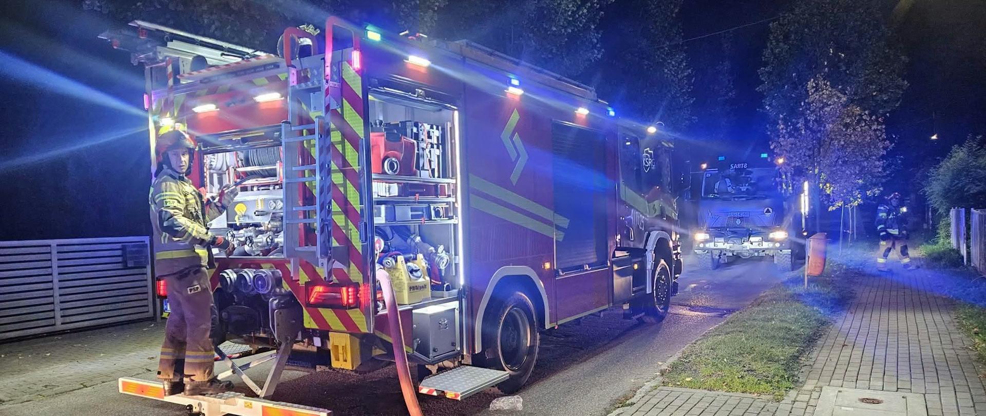 Na zdjęciu widoczny jest ciężki samochód ratowniczo-gaśniczy , należący do straży pożarnej, zaparkowany nocą na podjeździe lub bocznej drodze z włączonymi niebieskimi światłami sygnalizacyjnymi. Pojazd ma otwarte boczne i tylne skrytki sprzętowe, odsłaniające specjalistyczne wyposażenie techniczne oraz elementy autopompy. Strażak w pełnym umundurowaniu bojowym (w hełmie i ubraniu specjalnym) stoi na tylnym podeście pojazdu, obsługując sprzęt pożarniczy.