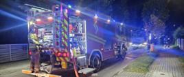 Na zdjęciu widoczny jest ciężki samochód ratowniczo-gaśniczy , należący do straży pożarnej, zaparkowany nocą na podjeździe lub bocznej drodze z włączonymi niebieskimi światłami sygnalizacyjnymi. Pojazd ma otwarte boczne i tylne skrytki sprzętowe, odsłaniające specjalistyczne wyposażenie techniczne oraz elementy autopompy. Strażak w pełnym umundurowaniu bojowym (w hełmie i ubraniu specjalnym) stoi na tylnym podeście pojazdu, obsługując sprzęt pożarniczy.