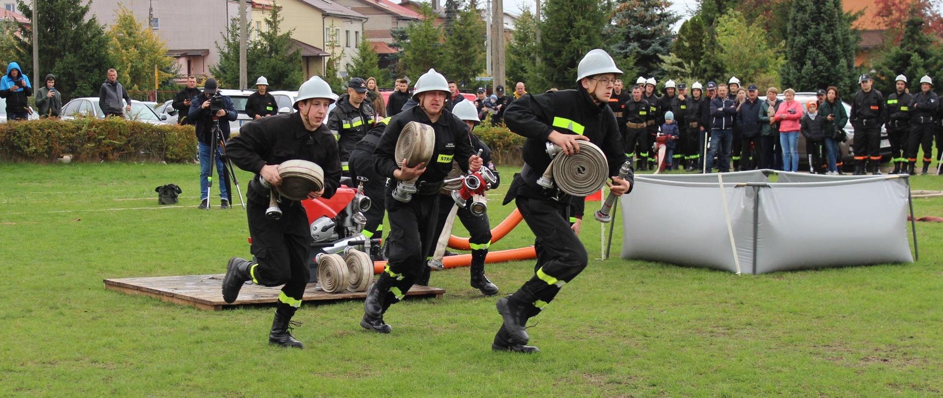 Czterech strażaków trzymających w dłoniach sprzęt pożarniczy rozpoczyna bieg w celu realizacji zadania – rozwinięcie bojowe. W tle widoczni są obserwatorzy zawodów sportowo-pożarniczych.