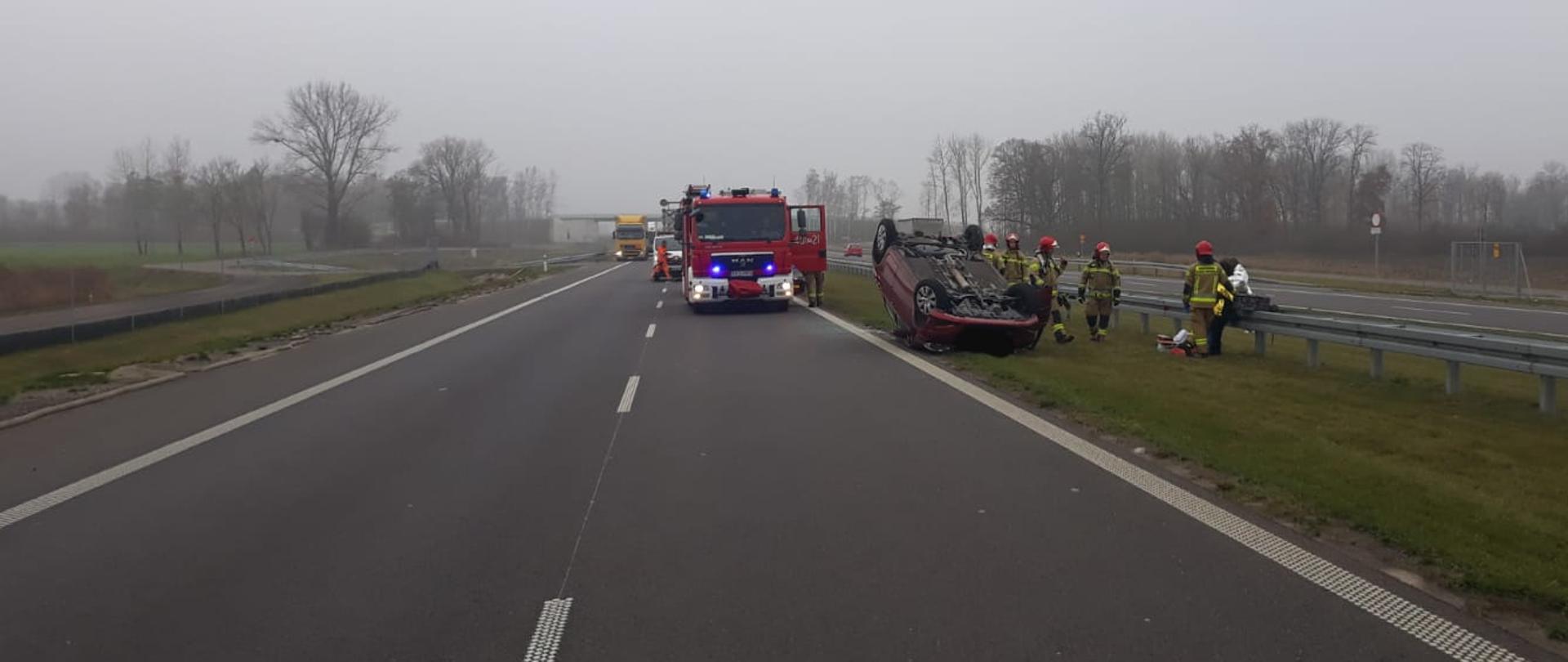 DACHOWANIE SAMOCHODU OSOBOWEGO NA DRODZE S5. Droga krajowa nr 5, na pasie zieleni na dachu leży samochód osobowy, obok stoją strażacy, samochód pożarniczy.
