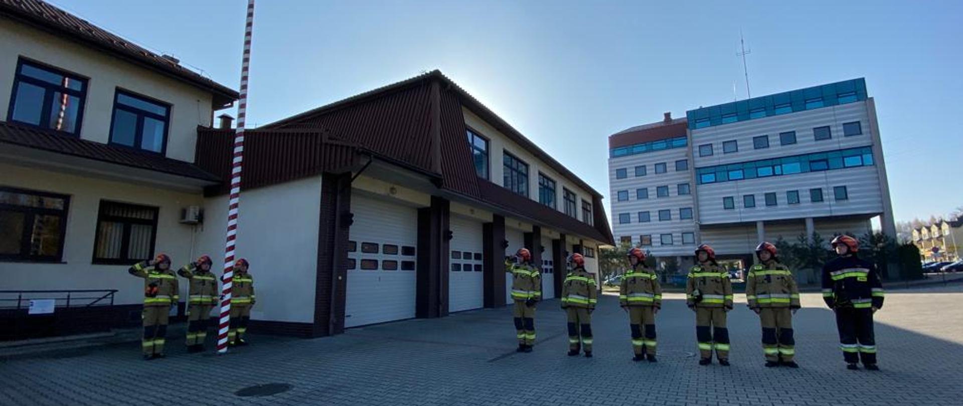 Na zdjęciu widoczni strażacy na tle budynku komendy. Na środku zdjęcia masz z flagą i salutujący trzej strażacy. Po prawej stronie zmiana służbowa w jednym szeregu z salutującym dowódcą zmiany służbowej.