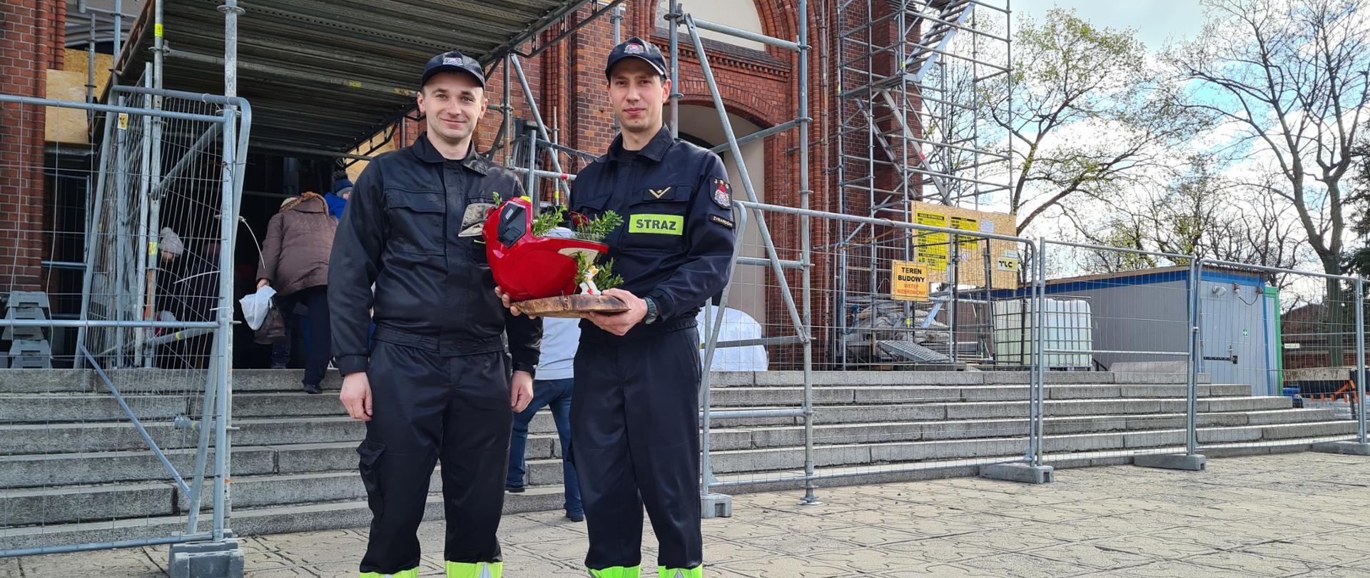 Dwóch strażaków stoi przed kościołem. Jeden z nich trzyma w rękach święconkę w hełmie