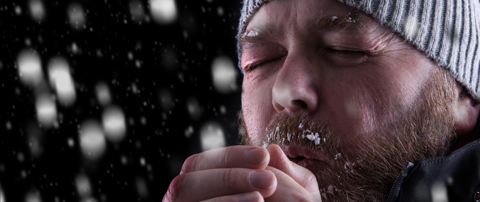 Marznący brodaty mężczyzna w czapce chucha na zmarznięte zaczerwienione ręce. Wokół jest ciemno i pada śnieg.