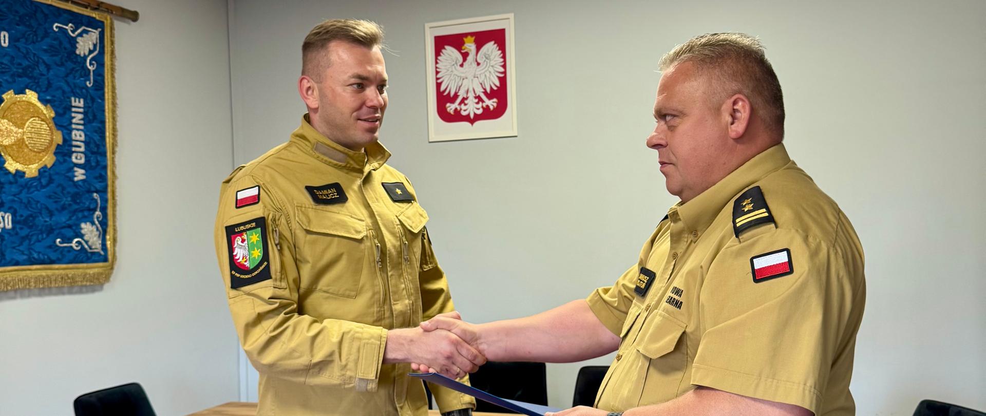 Komendant Powiatowy Państwowej Straży Pożarnej wręcza nagrodę funkcjonariuszowi podczas uroczystego spotkania służbowego. Obaj strażacy w umundurowaniu ściskają sobie dłonie. W tle widoczne są godło Rzeczypospolitej Polskiej oraz sala konferencyjna Komendy Powiatowej PSP.
